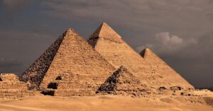 Three major Giza pyramids rising behind smaller desert ruins beneath dark clouds