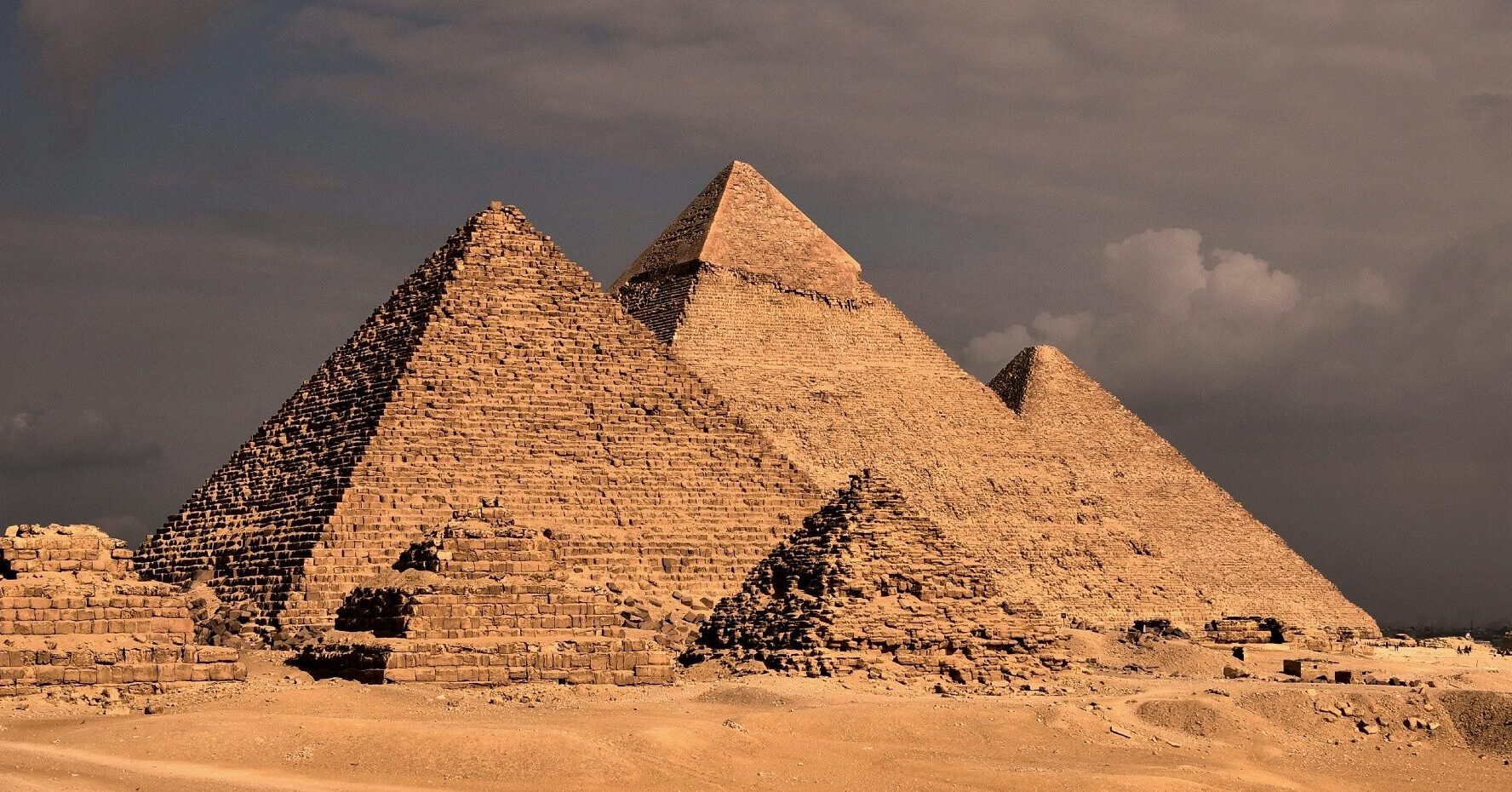 Three major Giza pyramids rising behind smaller desert ruins beneath dark clouds