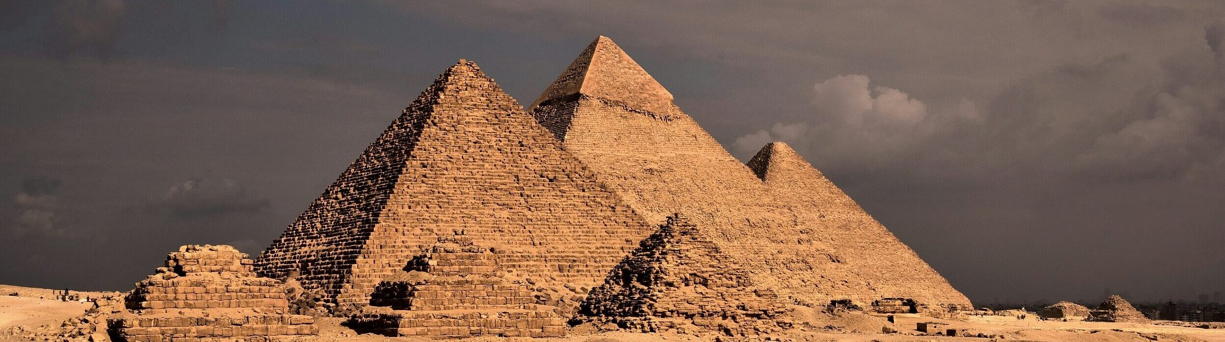 Three major Giza pyramids rising behind smaller desert ruins beneath dark clouds