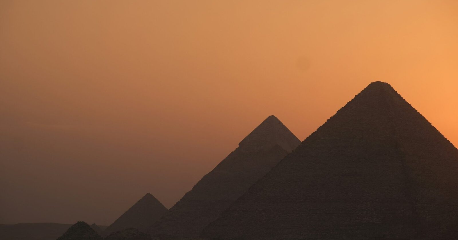 Three Giza pyramids silhouetted in overlapping perspective against a hazy orange sunset sky