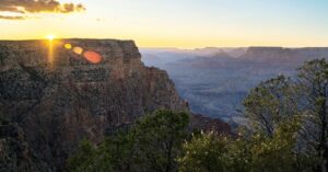 Sunset starburst at the edge of a Grand Canyon rim cliff with trees in the foreground and hazy canyon layers beyond
