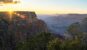 Sunset starburst at the edge of a Grand Canyon rim cliff with trees in the foreground and hazy canyon layers beyond