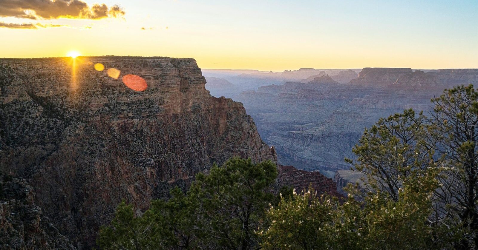 Sunset starburst at the edge of a Grand Canyon rim cliff with trees in the foreground and hazy canyon layers beyond