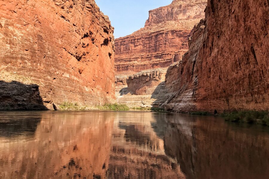 Red Grand Canyon cliffs reflected in calm Colorado River water beneath a clear blue sky
