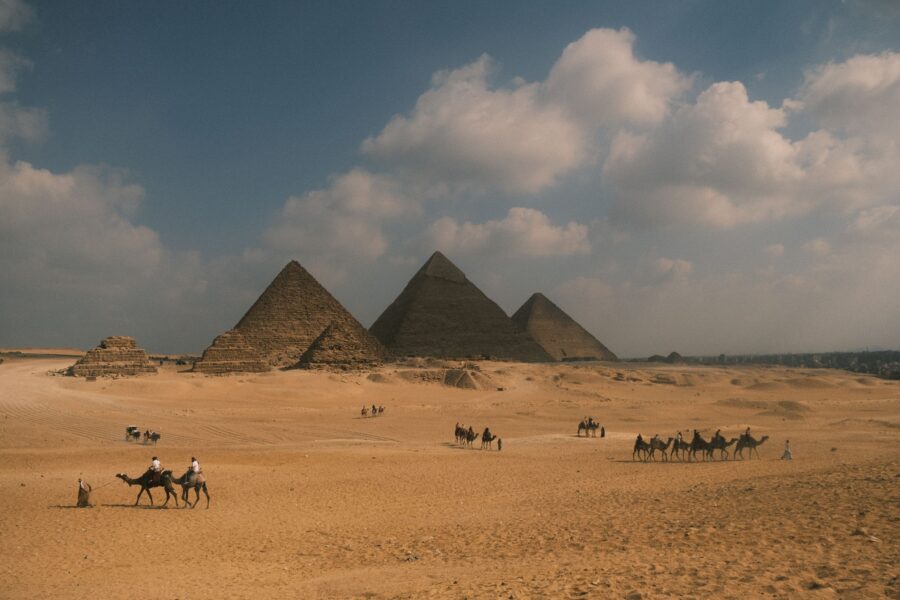 Camel riders crossing the Giza Plateau with the three major pyramids beneath a broad blue sky